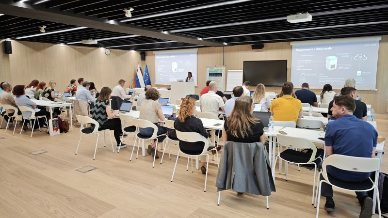 EDIAQI consortium members seated in a modern conference room at the National Institute of Biology in Ljubljana during a project meeting. A presenter stands at a podium in front of two large screens displaying slides about sensors and data transfer. Flags of Slovenia and the EU are visible near the podium.