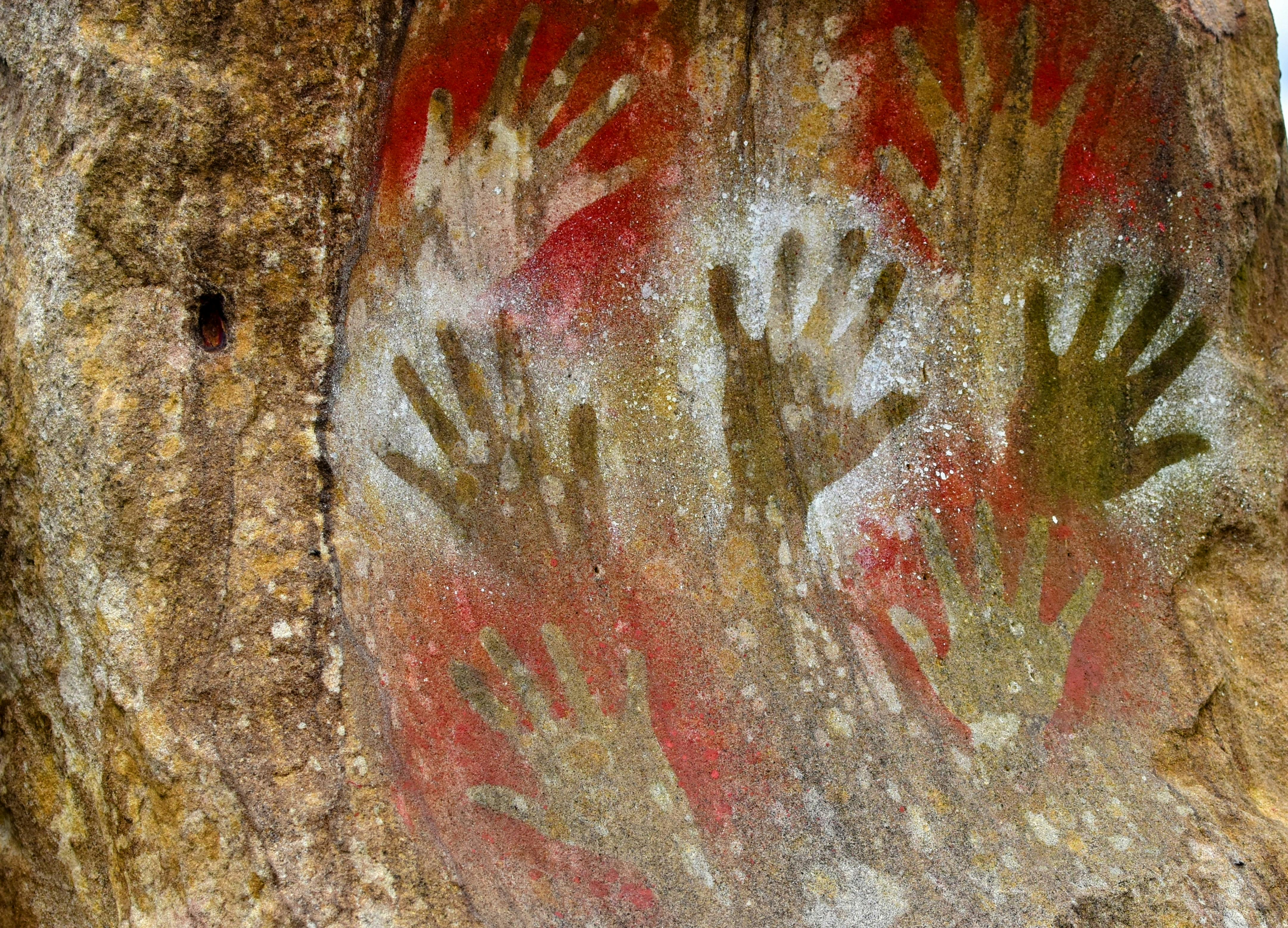 Ancient cave painting showing multiple stenciled handprints in red, white, and yellow pigments on a rough rock surface.