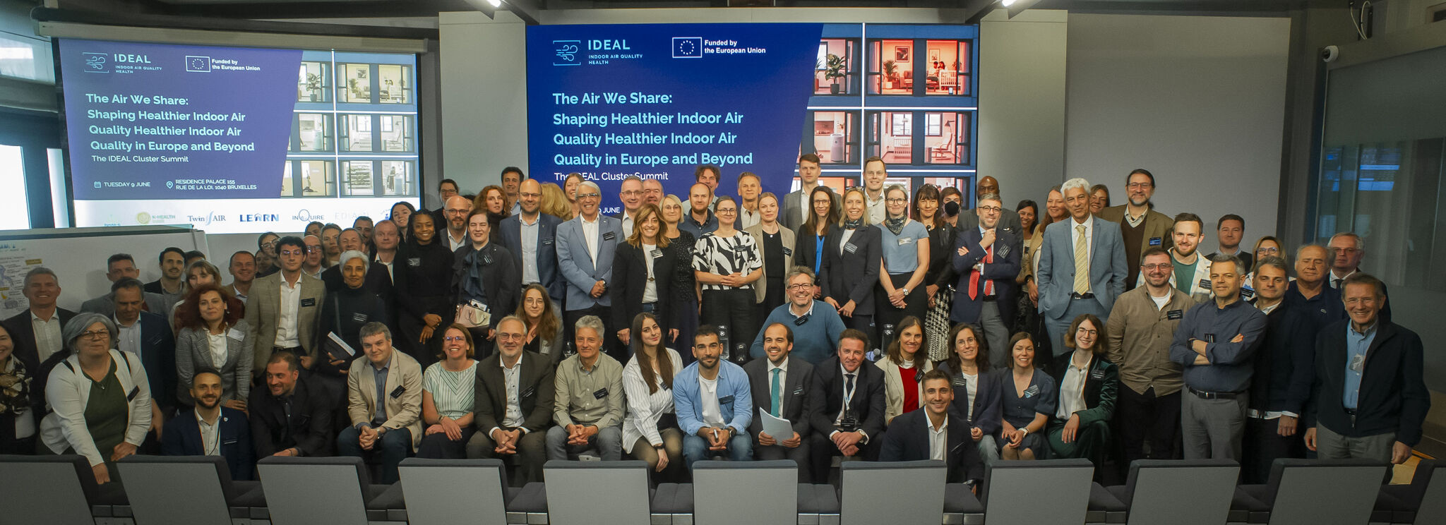 Participants gather for a group photograph at the IDEAL Cluster Summit – The Air We Share: Shaping Healthier Indoor Air Quality in Europe and Beyond in Brussels. Researchers, policymakers, industry representatives, public authorities and civil society organisations from across Europe stand together in front of the Summit stage, representing the seven Horizon Europe projects of the IDEAL Cluster and celebrating their shared commitment to advancing healthier indoor environments through research, innovation an