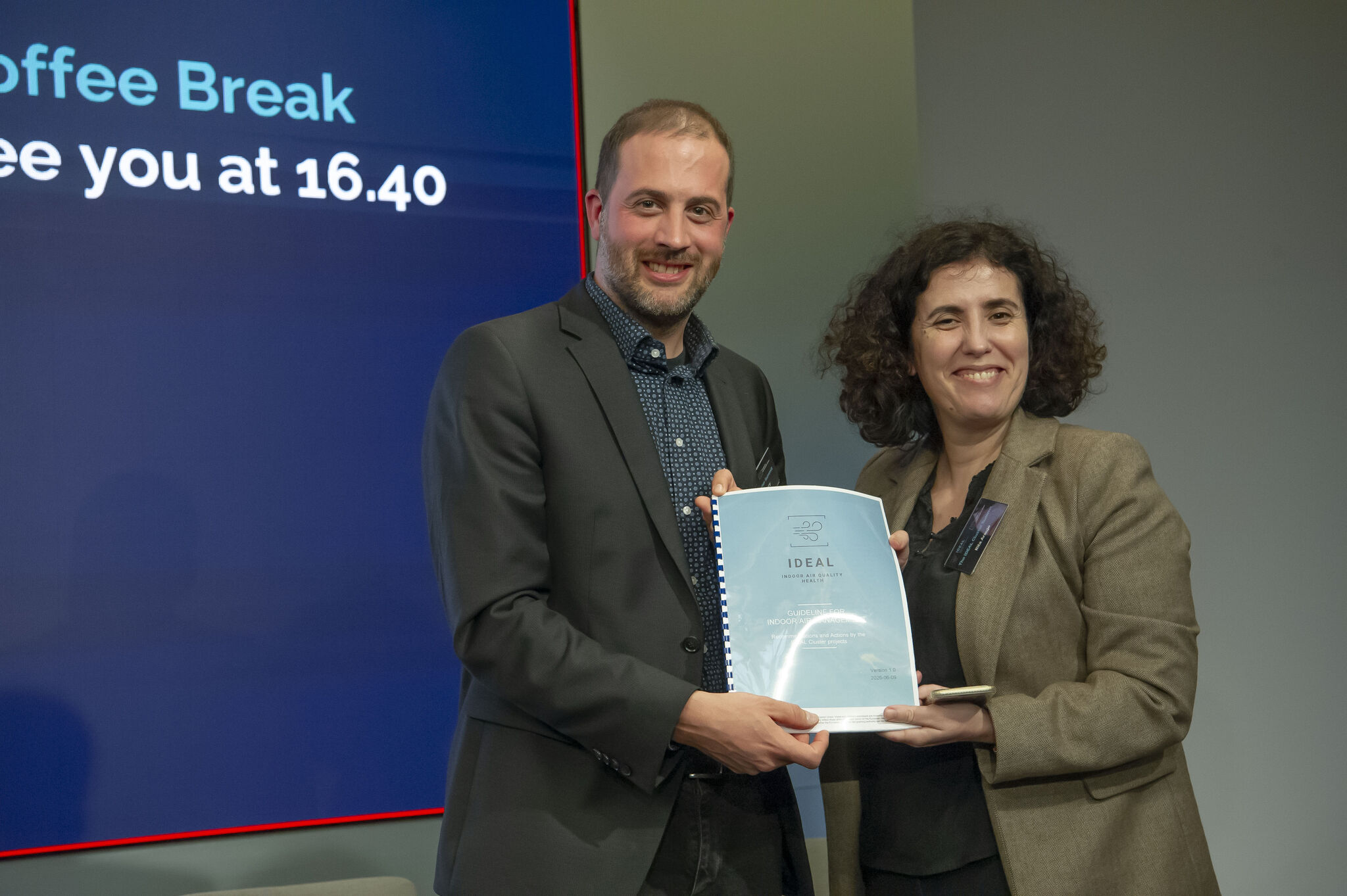 Christian Goroncy, Team Manager at the German Institute for Standardisation (DIN), presents the IDEAL Cluster Indoor Air Quality Guidelines to Rita Araujo, Policy Officer at the European Commission's Directorate-General for Research and Innovation (DG RTD), during the IDEAL Cluster Summit – The Air We Share in Brussels. The handover symbolises the IDEAL Cluster's commitment to translating collaborative research into practical guidance that can support future European policy and healthier indoor environments.