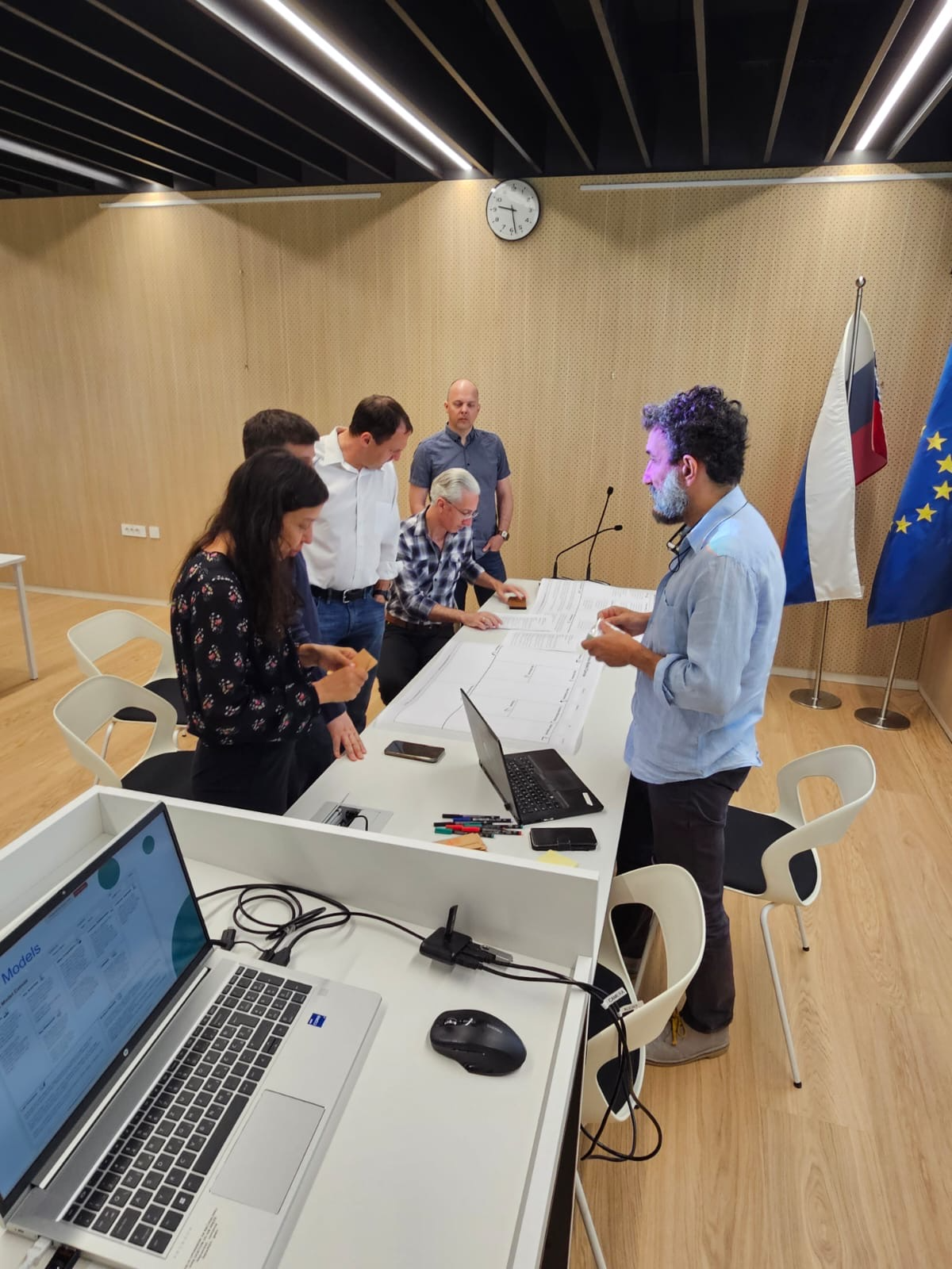 EDIAQI consortium members participating in a business model canvas exercise during the project meeting at the National Institute of Biology in Ljubljana. Six people are gathered around a table discussing and pointing at a large printed canvas, with laptops and markers visible. Slovenian and EU flags stand in the background.