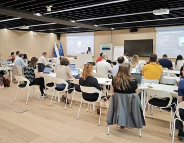 EDIAQI consortium members seated in a modern conference room at the National Institute of Biology in Ljubljana during a project meeting. A presenter stands at a podium in front of two large screens displaying slides about sensors and data transfer. Flags of Slovenia and the EU are visible near the podium.