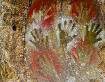 Ancient cave painting showing multiple stenciled handprints in red, white, and yellow pigments on a rough rock surface.